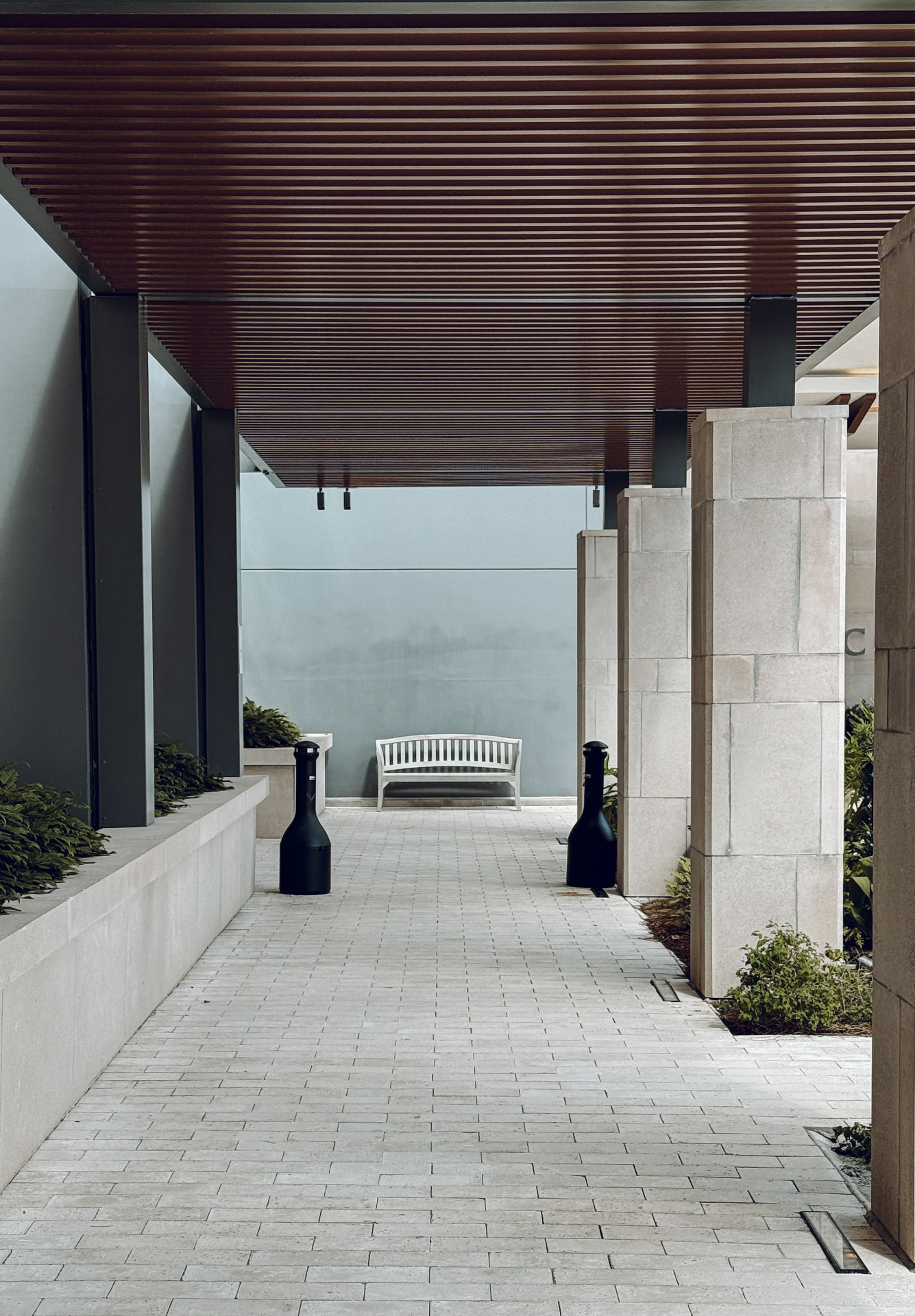 Passagem externa coberta com piso claro de peças alongadas, jardineiras laterais e banco ao fundo